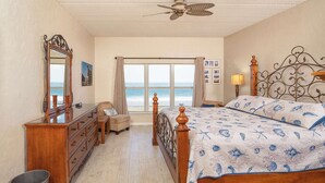 Een slaapkamer met een houten bed, een dressoir, een spiegel, een plafondventilator en uitzicht op de zee door grote ramen.