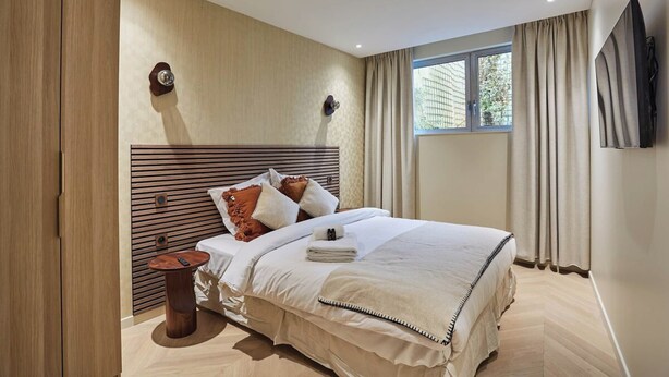 A modern hotel room with a large bed, a wooden headboard, a small side table, and a large window with curtains. - Elegant Apartment St Germain des Pres Luxembourg Park (Paris)
