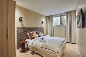 Une chambre d’hôtel moderne avec un grand lit, une tête de lit en bois, une petite table de chevet et une grande fenêtre avec des rideaux.