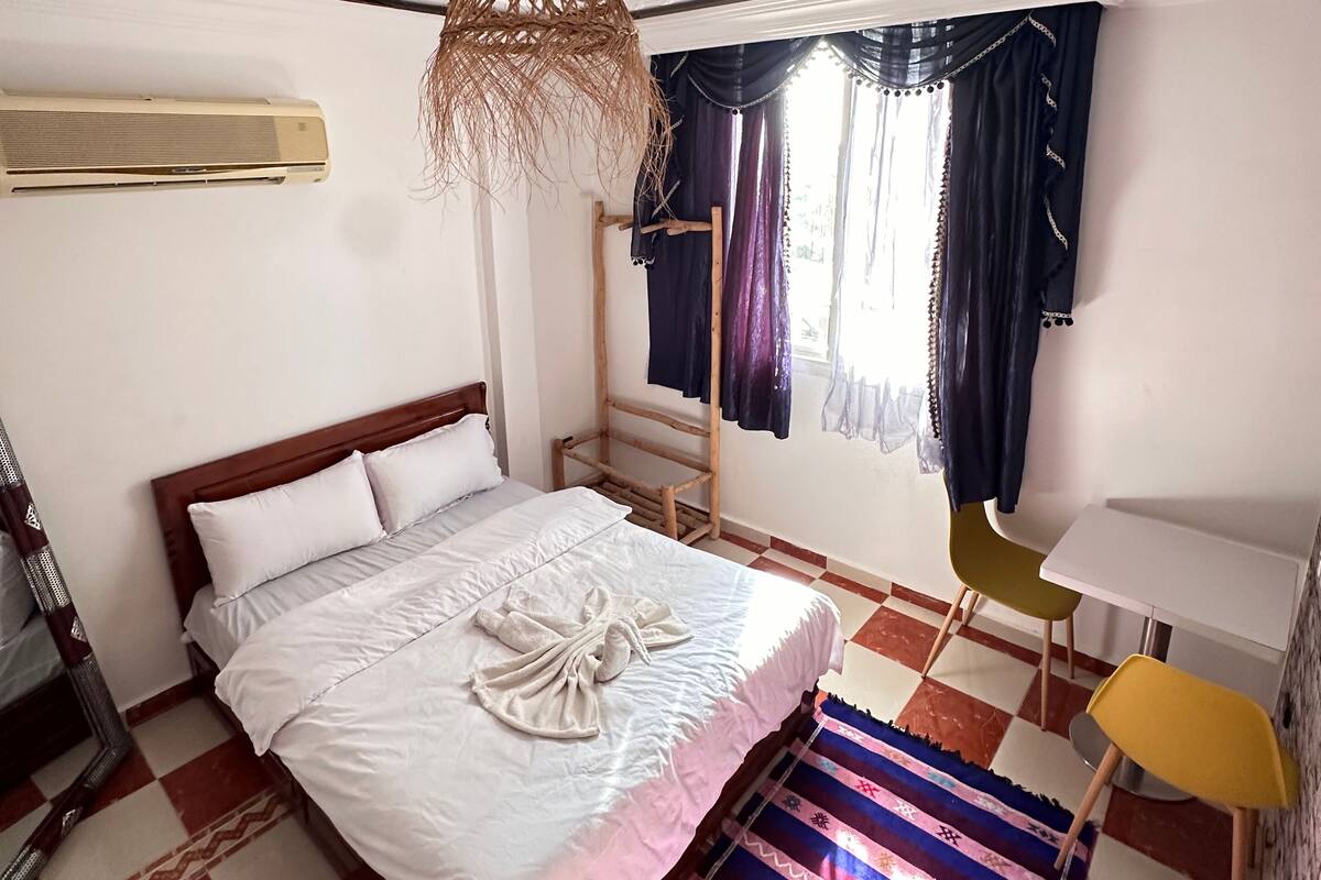 A bedroom with a bed, a hanging light fixture, a window with curtains, a chair, and a small table.