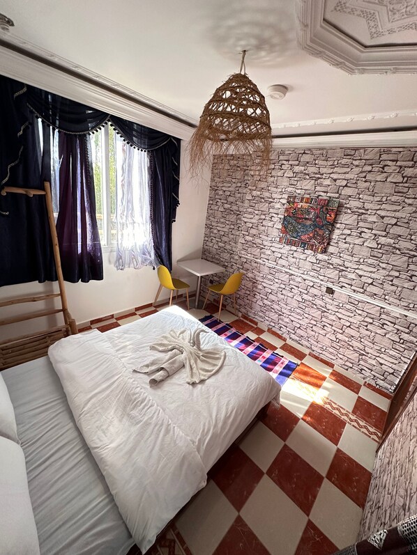 A bedroom with a bed, a wooden ladder, a small table, two yellow chairs, a woven hanging light fixture, and a patterned wall. - sunrise auberge (Aourir)