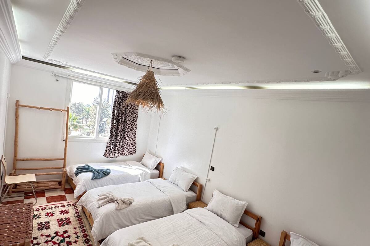 A room with three beds, a patterned rug, a hanging light fixture, and a window with a view.