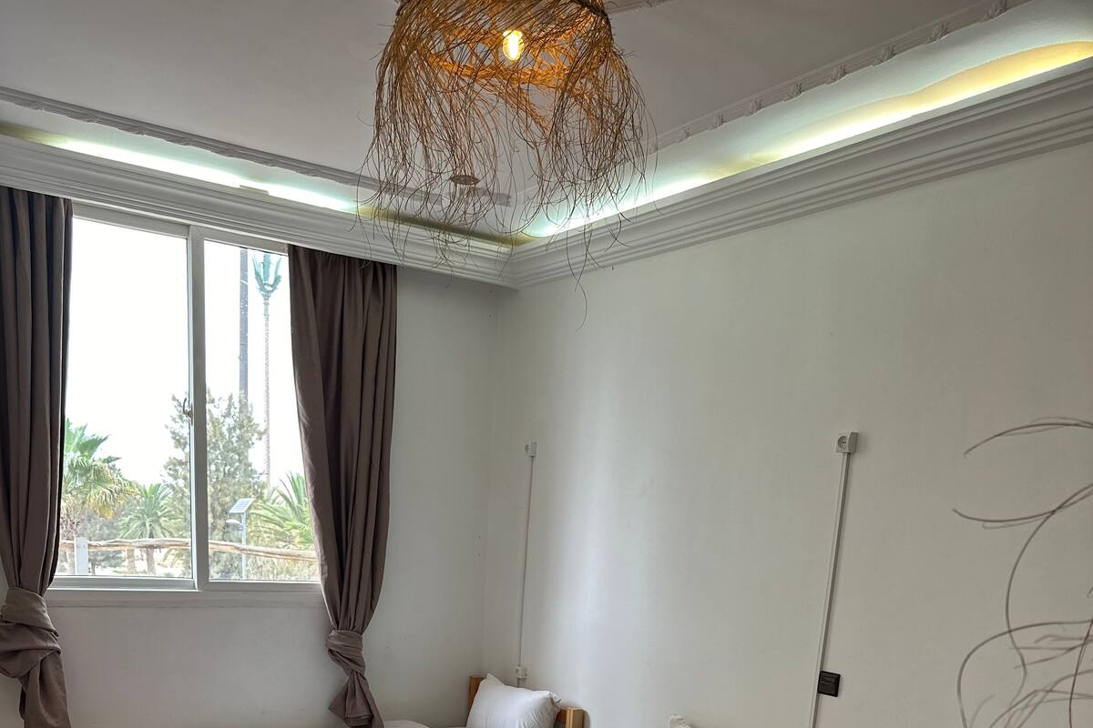 A hotel room with three beds, a checkered floor, a hanging light fixture, and a view of palm trees through the window.