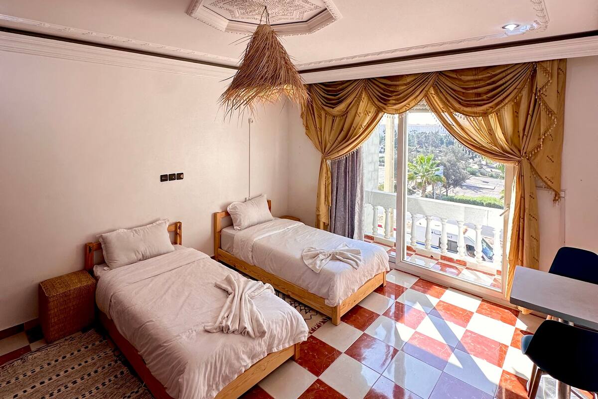 A hotel room with two beds, a checkered floor, a balcony with curtains, and a hanging light fixture.