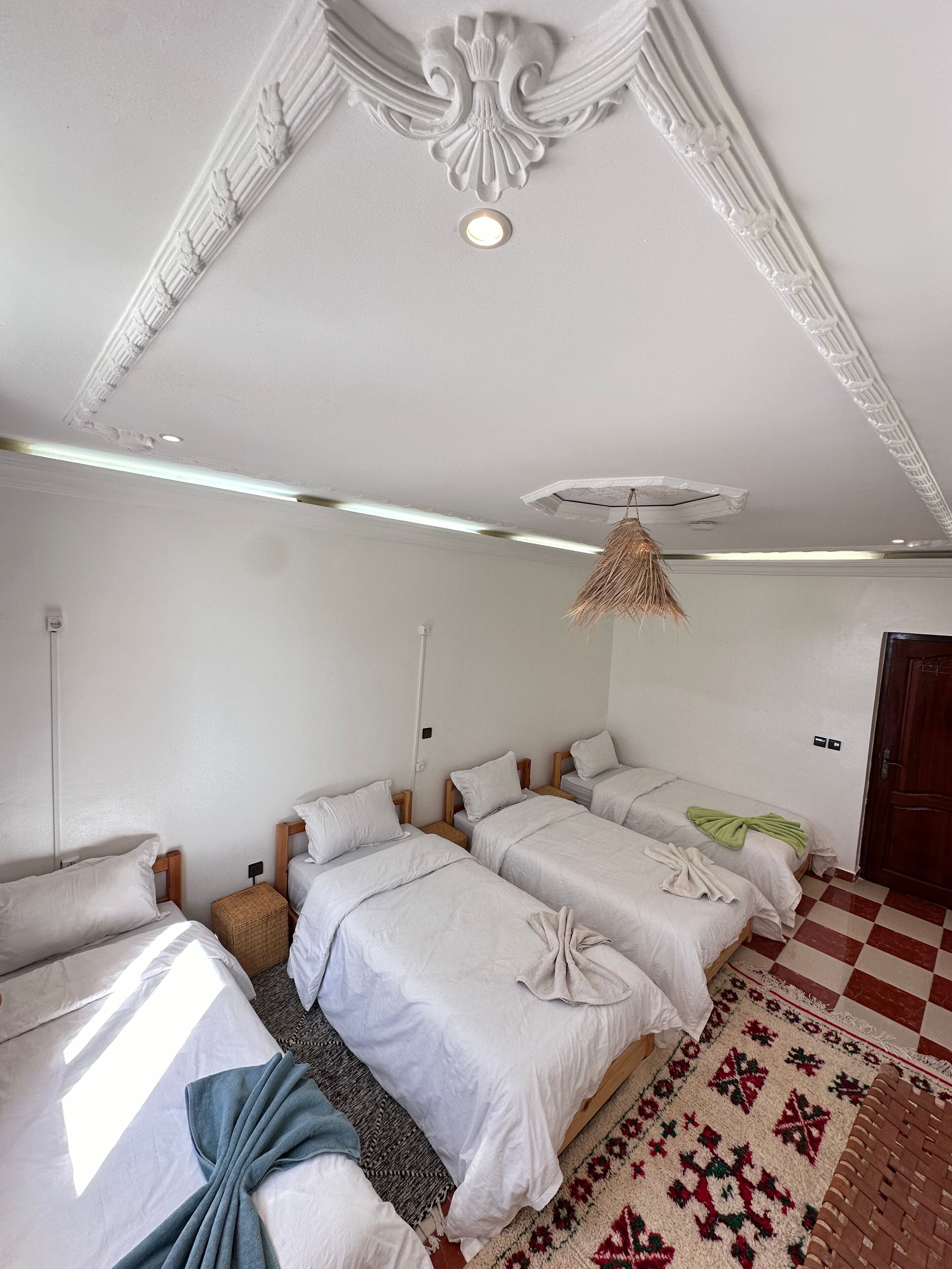 Une chambre d’hôtel avec trois lits, une tête de lit en osier, un tapis à motifs et un plafond orné de moulures décoratives.