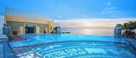 An infinity pool with a view of the sunset over the water, a modern building with a patterned facade, and a stone statue on the right.