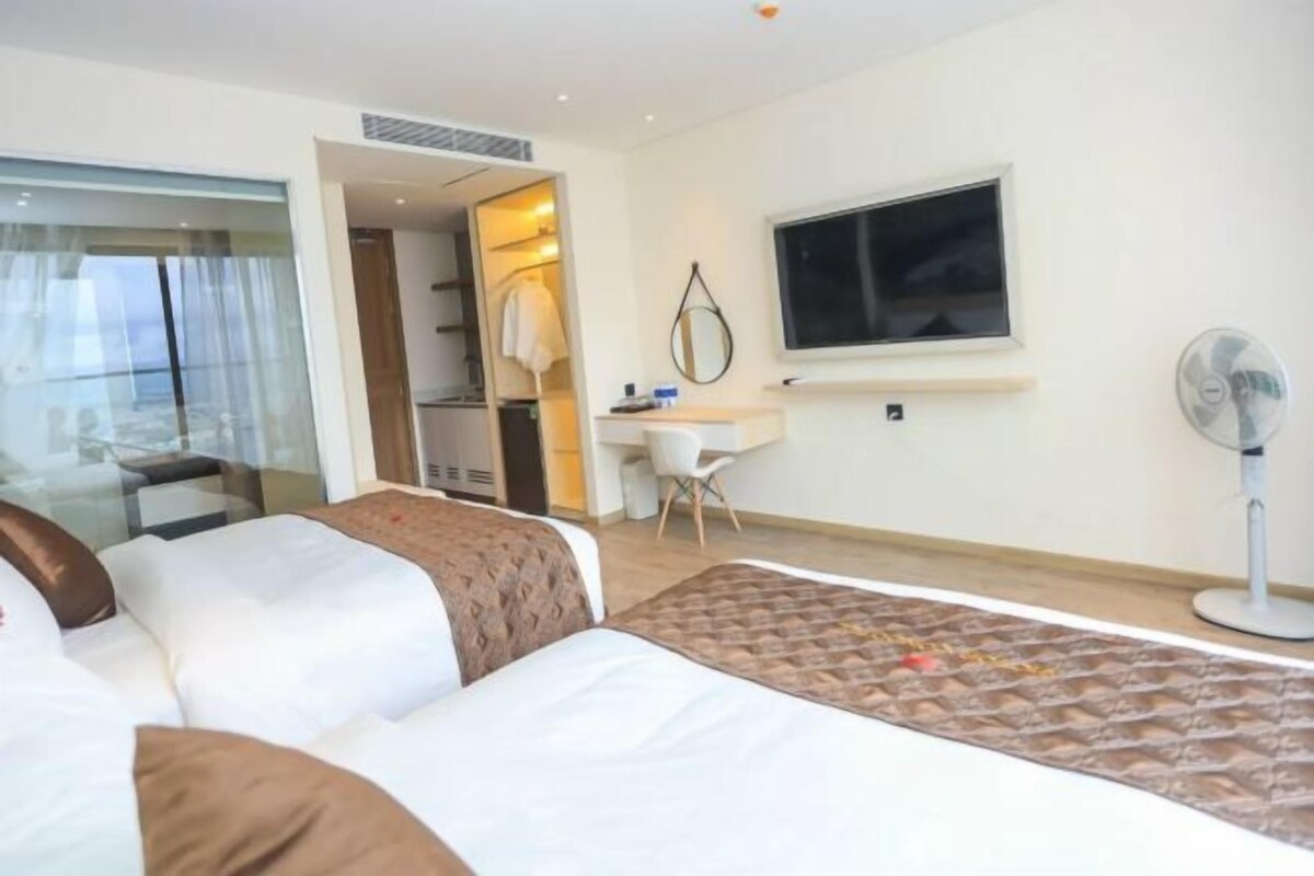 A hotel room with two beds, a desk, a chair, a TV, and a fan.
