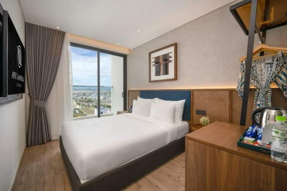 A hotel room with a large bed, a desk, a view of the city, and a framed picture on the wall.