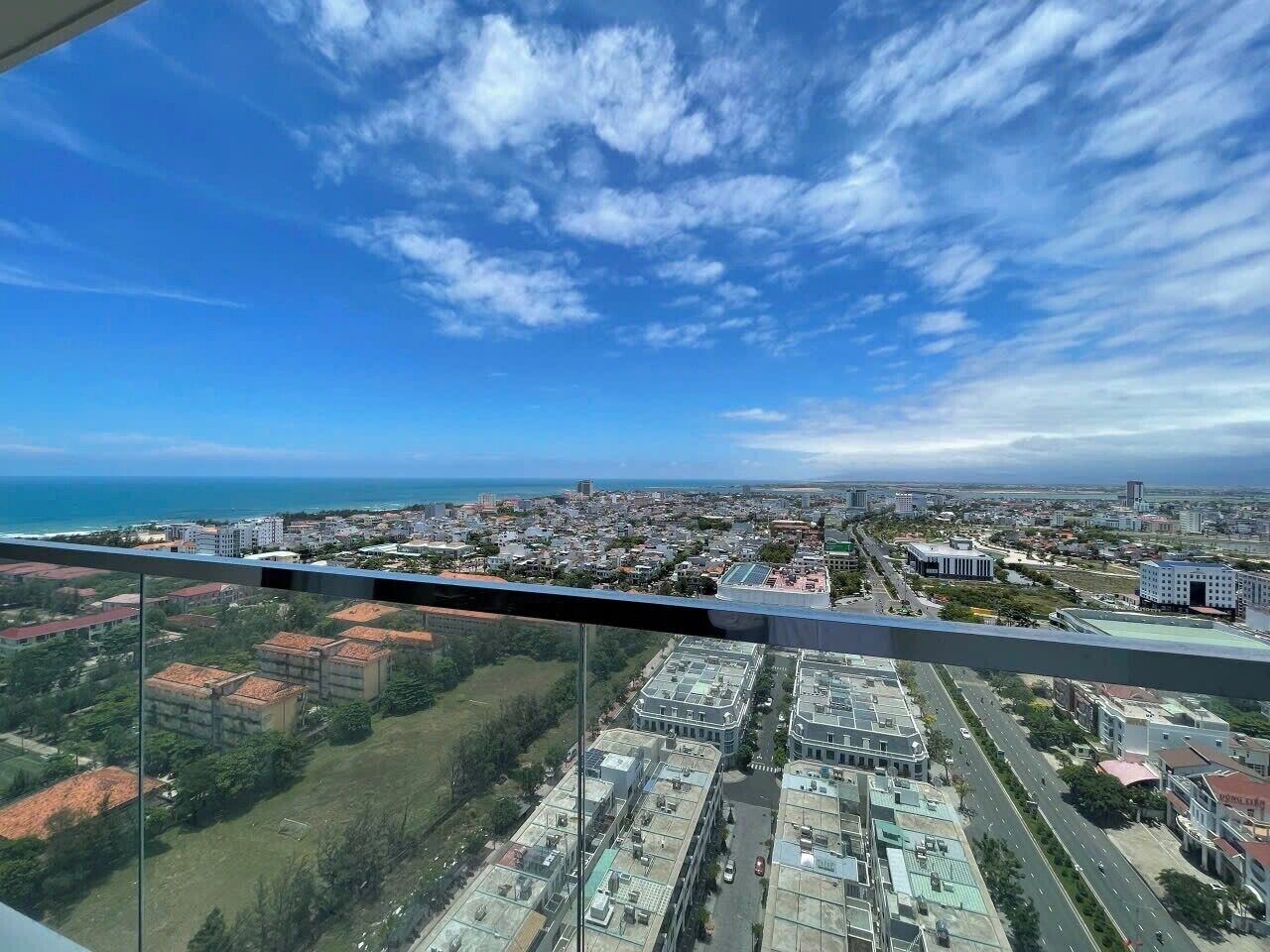 A panoramic view of a cityscape from a high vantage point, featuring a mix of residential and commercial buildings, a clear blue sky, and a distant coastline.