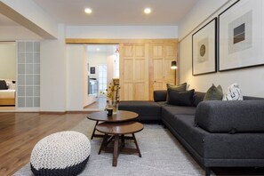 A modern living room with a grey sectional sofa, a round coffee table, and a white pouf. There is a wooden door and a framed picture on the wall.