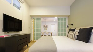 A hotel room with a bed, a TV on a black cabinet, a desk with chairs, and a door leading to another room.