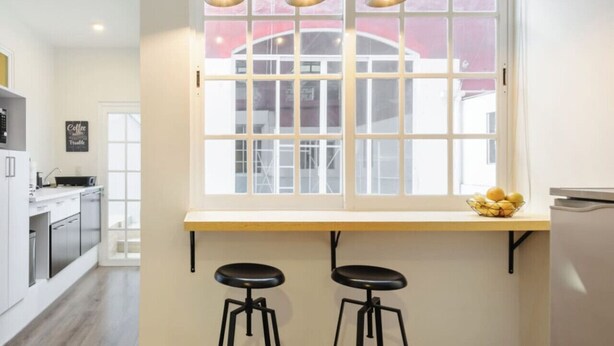 A modern kitchen with a large window, two bar stools, and hanging pendant lights.
