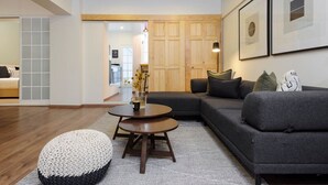 A modern living room with a grey sectional sofa, a round coffee table, and a white pouf. There is a wooden door and a framed picture on the wall.