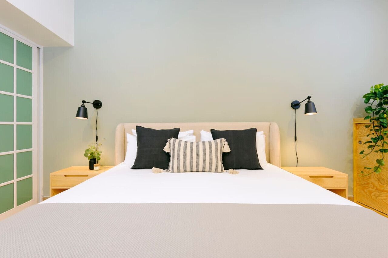 A neatly made bed with a white duvet, two black pillows, and a striped pillow. There are two bedside tables with lamps, a small plant, and a wooden cabinet with a plant.