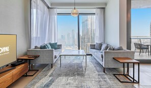 Ein modernes Wohnzimmer mit einer Sofagarnitur, einem Couchtisch und einem Fernseher auf einer Holzkonsole. Große Fenster bieten Blick auf die Stadt.