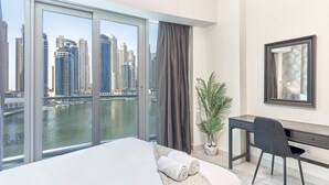 Ein modernes Hotelzimmer mit großem Fenster und Blick auf die Stadt, einem Bett mit aufgerollten Handtüchern, einem Schreibtisch, einem Stuhl und einer Topfpflanze.