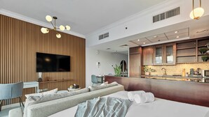 A modern hotel room with a large bed, a seating area with a sofa and armchairs, a flat-screen TV mounted on a wooden wall, a kitchenette with a microwave, and a ceiling fan. - bnbme | 8 Boulevard Walk | Studio (Dubai)