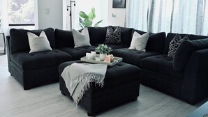 A modern living room with a black sectional sofa, a grey ottoman, and a small side table with candles and a plant. - Private Pickleball Oasis + Pool + 15 Min to Beaches & El Yunque, 30 Min Airport (Río Grande)