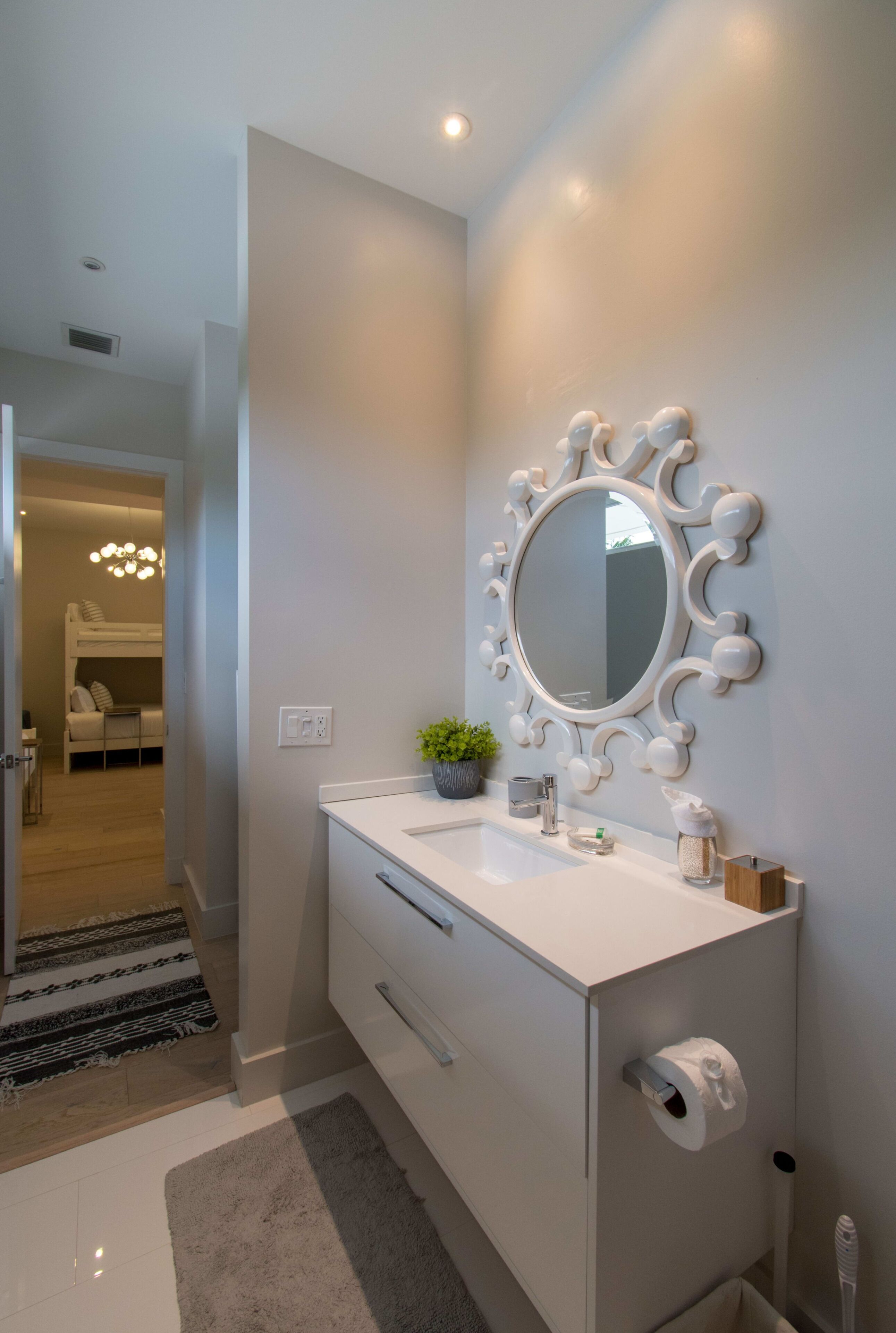 Un baño moderno con un tocador blanco, un espejo redondo con un marco decorativo, una planta pequeña en la encimera y un dispensador de papel higiénico.