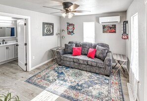 Un salón con un sofá gris, cojines rojos, un tapete estampado, un ventilador de techo y decoración de la Ruta 66.