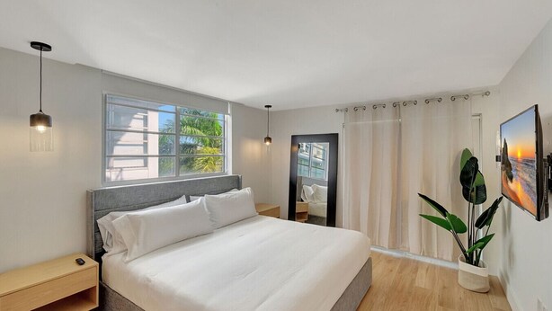A modern hotel room with a large bed, a bedside table, a nightstand, a window with a view of greenery, a mirror, a TV, and a potted plant. - Modern 2-Bedroom in Central South Beach Location (Miami Beach)