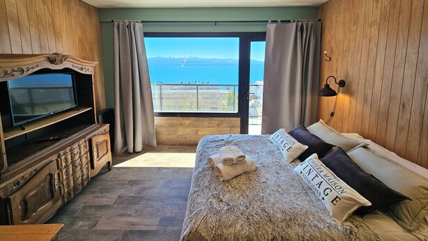 Une chambre dotée d’un grand lit, d’une télévision, de lambris en bois, d’un balcon avec vue sur la mer et d’un luminaire au plafond.