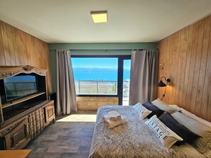 Ein Schlafzimmer mit einem großen Bett, einem Fernseher, Holzvertäfelung, einem Balkon mit Meerblick und einer Deckenleuchte.
