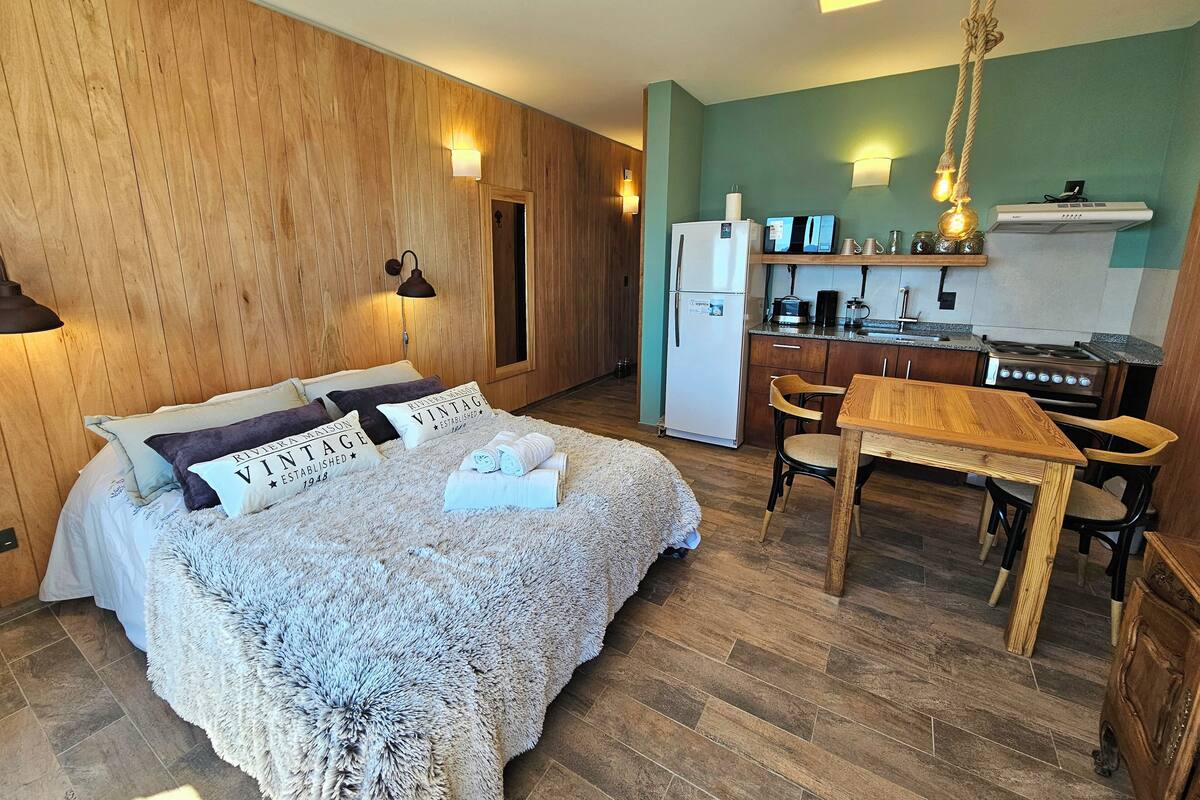 Una habitación acogedora con cama, cabecero de madera, kitchenette con refrigerador, estufa y mesa de comedor con sillas.