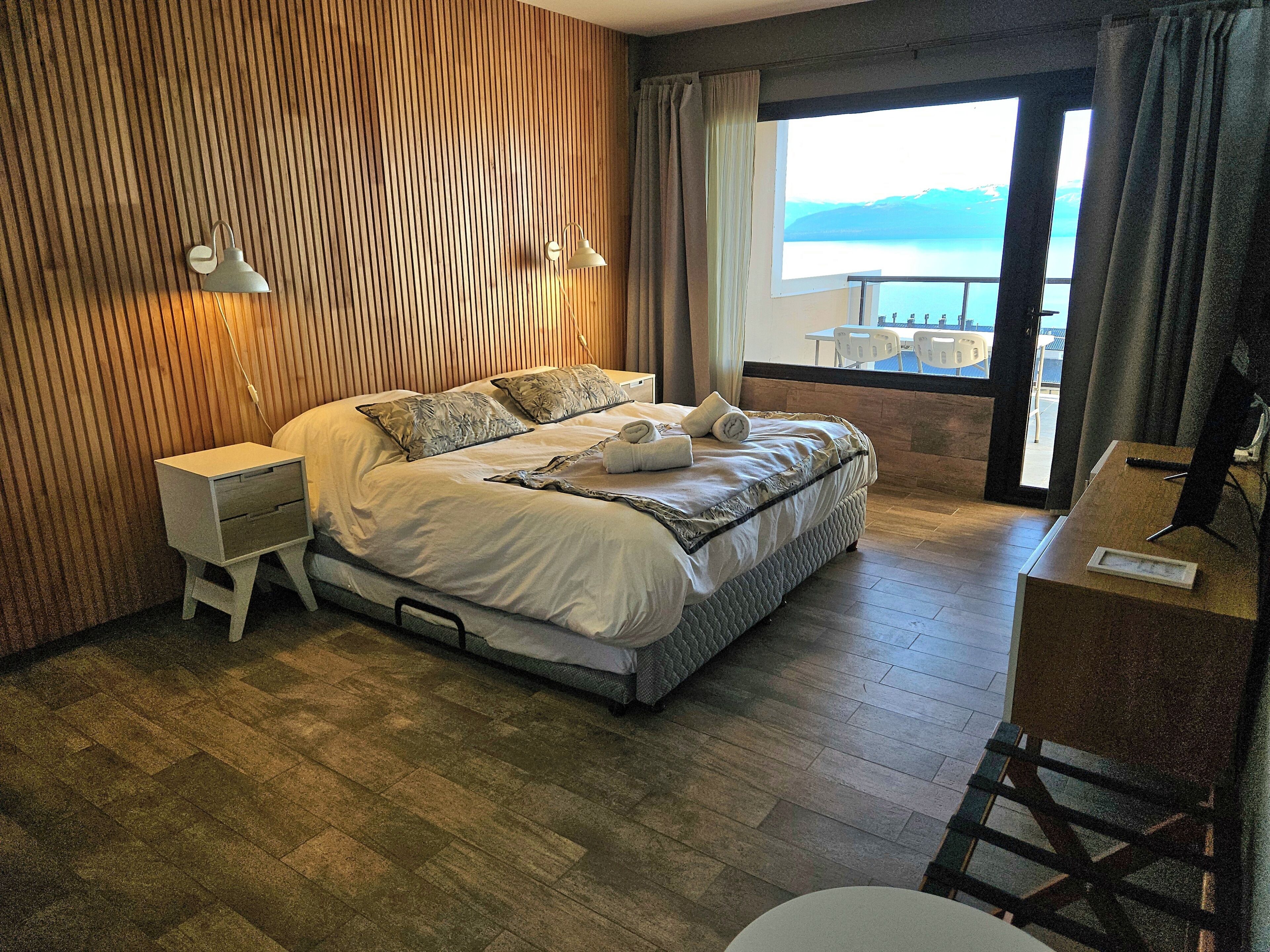 A hotel room with a bed, bedside tables, a desk, and a view of the sea.