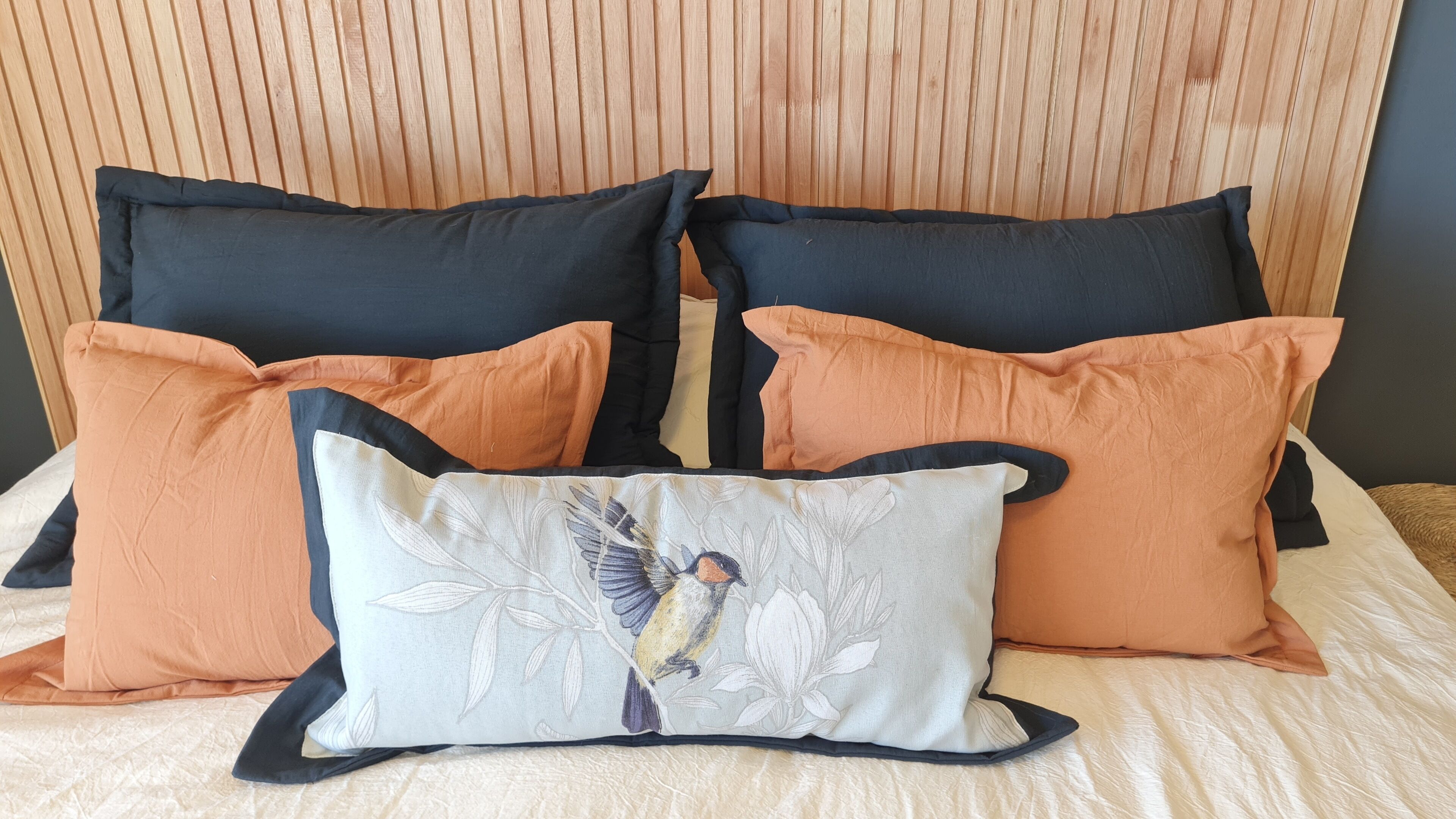 Una cama con cabecera de madera, varios cojines en negro y naranja, y un cojín decorativo con diseño de pájaro.