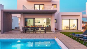 A modern two-story house with a swimming pool, outdoor dining area, and a well-lit interior.