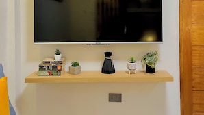 Un téléviseur à écran plat fixé au mur, au-dessus d’une étagère en bois où sont posés des livres et de petites plantes en pot.