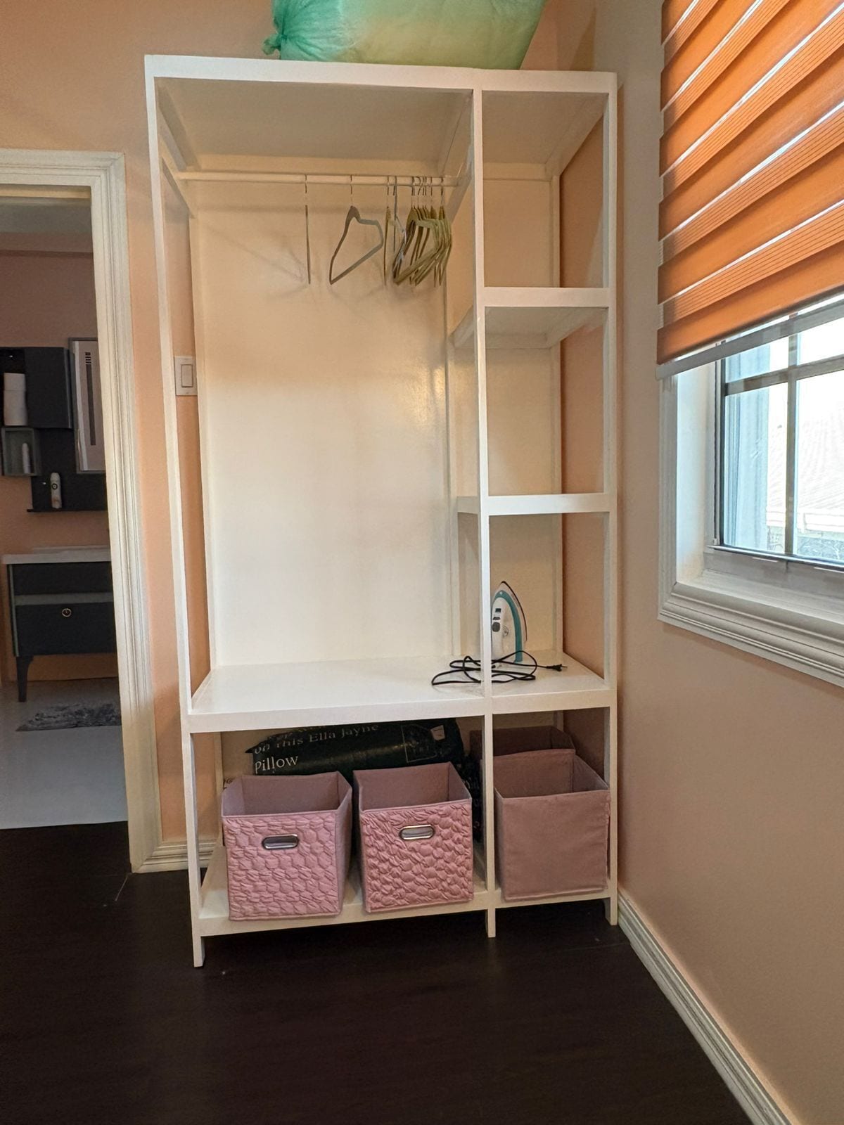 A white shelving unit with pink storage bins, a hanging rack, and a window with blinds.