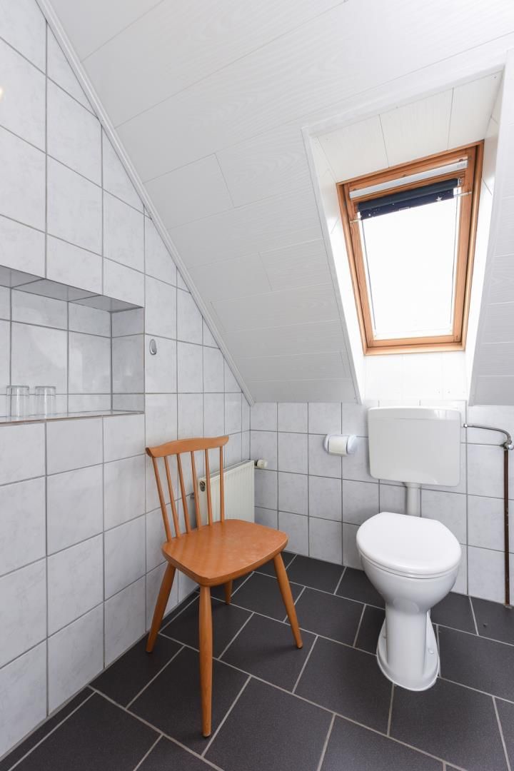 Ein kleines, modernes Badezimmer mit schrägem Dach, einem Oberlicht, einer weißen Toilette und einem Holzstuhl.