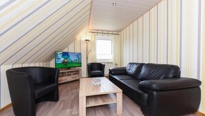 A cozy living room with a wooden coffee table, black leather sofa, and armchairs. A flat-screen TV is mounted on the wall, and a window provides natural light. - House Hafeneck FeWo Deichblick (Bensersiel)