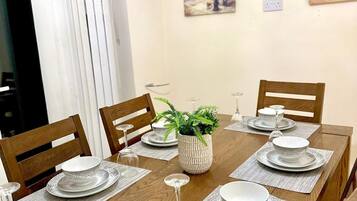 A dining room with a long wooden table set for six, surrounded by wooden chairs. There are two paintings on the wall, a potted plant on the table, and a window with white curtains.