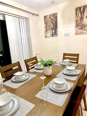 Een eetkamer met een lange houten tafel voor zes personen, omringd door houten stoelen. Twee schilderijen hangen aan de muur, op de tafel staat een potplant en er is een raam met witte gordijnen.