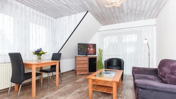 A modern living room with a wooden dining table, black chairs, a purple sofa, a glass coffee table, a TV on a wooden cabinet, and sheer curtains.
