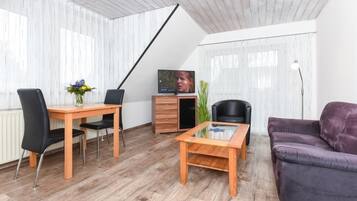 Una sala de estar moderna con una mesa de comedor de madera, sillas negras, un sofá púrpura, una mesa de centro de vidrio y un televisor sobre un mueble de madera, además de cortinas de gasa.
