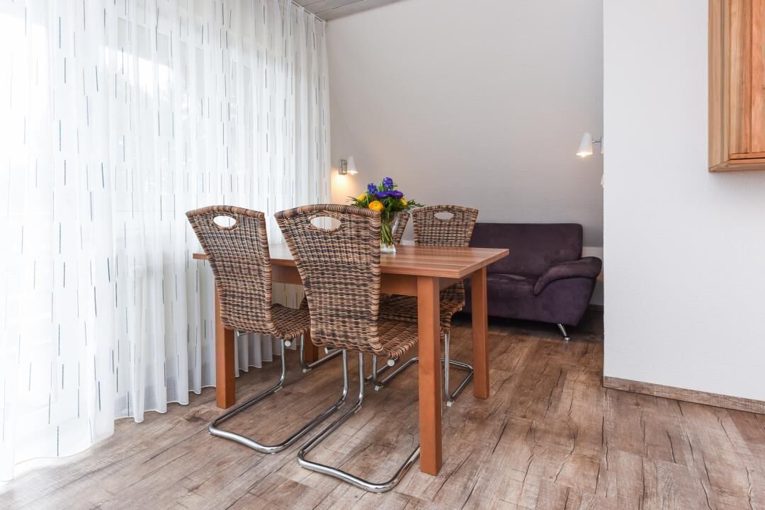 Ein Essbereich mit einem Holztisch, Korbstühlen, einem violetten Sofa und einem weißen Vorhang.