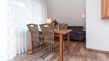 Ein Essbereich mit einem Holztisch, Korbstühlen, einem violetten Sofa und einem weißen Vorhang.