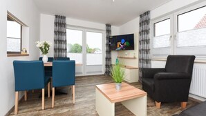 A modern living room with a dining area, a black armchair, a TV, and a plant on a wooden table. - Haus zur Möwe apartment Möwe 1 (Bensersiel)