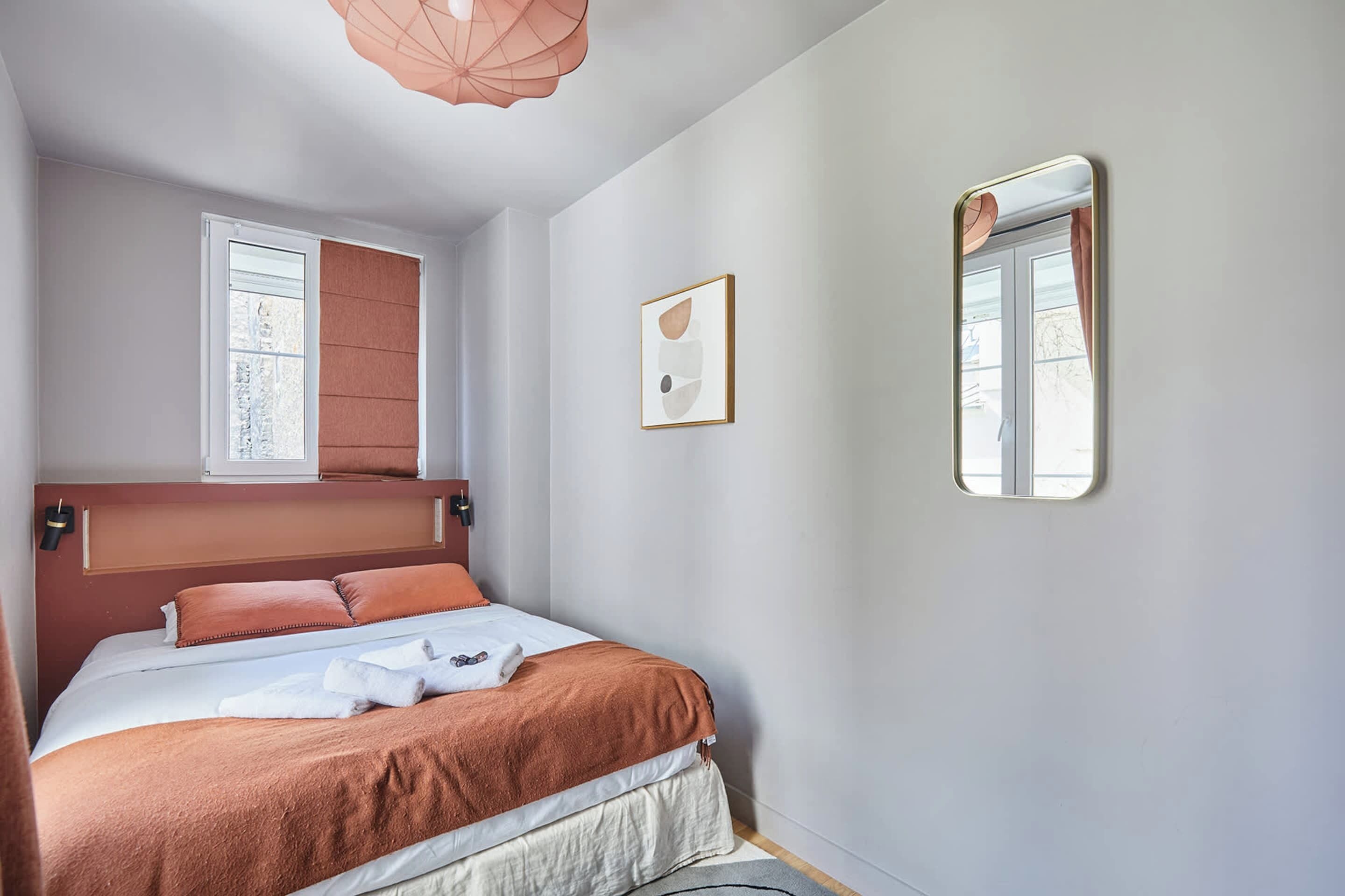 Ein Schlafzimmer mit Bett, Fenster, Wandspiegel und Deckenleuchte.