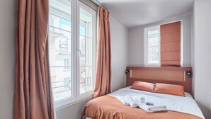Uma cama bem arrumada com cabeceira marrom, uma janela com cortinas, e vista para edifícios do lado de fora.
