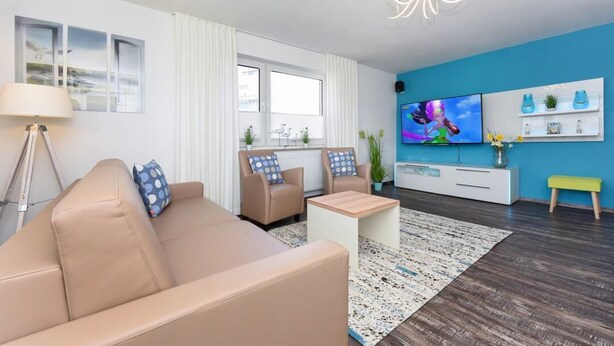 A modern living room with a beige sofa, two armchairs, a coffee table, and a TV on a blue wall. - Haus zur Möwe apartment Möwe 2 (Bensersiel)