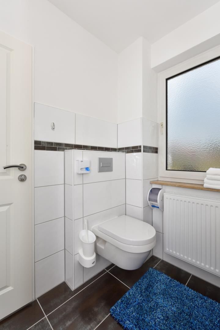 A modern bathroom with white tiles, a toilet, a hand dryer, and a blue rug.
