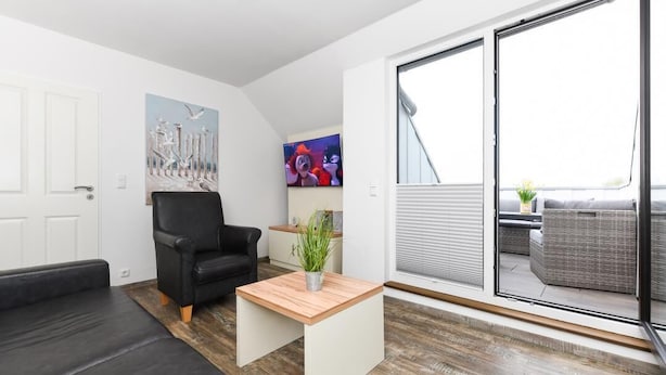 A modern living room with a black leather sofa, a wooden coffee table, and a black armchair. There is a sliding glass door leading to an outdoor area with wicker furniture. - Haus zur Möwe apartment Möwe 4 (Bensersiel)