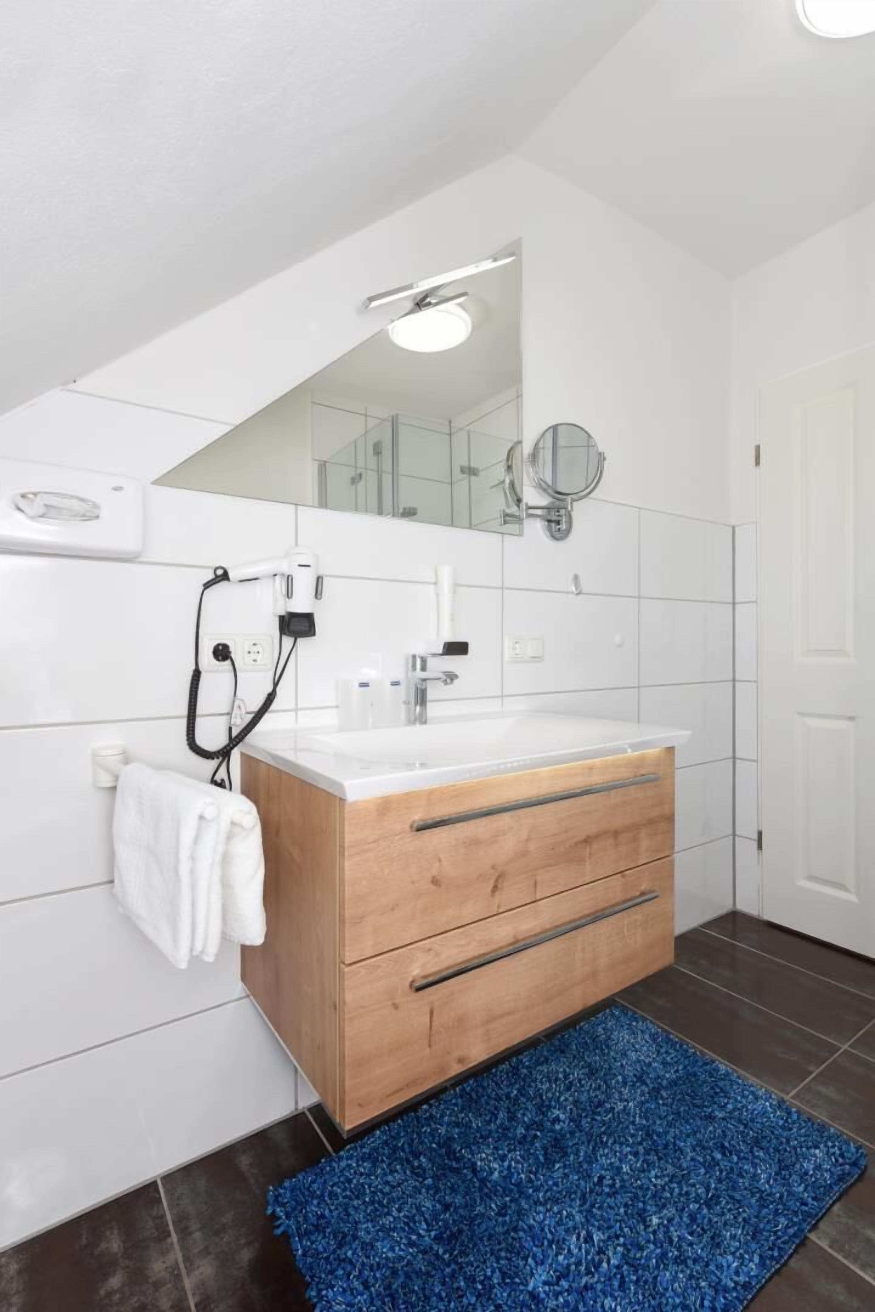 Ein modernes Badezimmer mit einem Holzlagerfläschchen, weißen Fliesen, einem blauen Teppich und einem Spiegel mit rundem Rahmen.