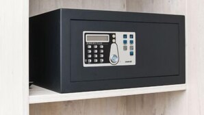 A black safe with a keypad and a fingerprint scanner, mounted on a white shelf against a wooden background. - House Strandkorb apartment Strandkob 2 (Bensersiel)