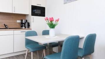 Une cuisine moderne avec des meubles blancs, un four à micro-ondes et une machine à café. On y trouve une table à manger avec des chaises bleues et un vase rempli de fleurs roses.